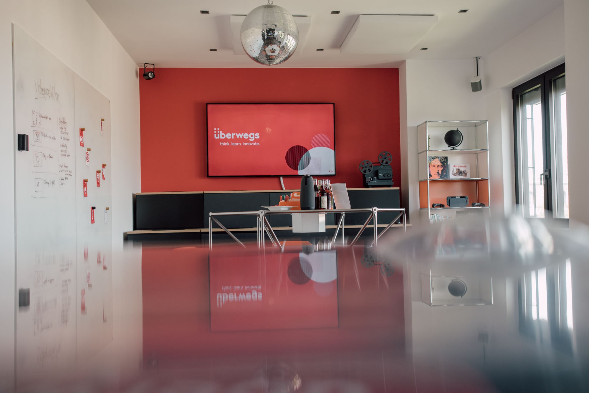 Das Bild zeigt einen modernen Besprechungsraum mit einem roten Hintergrund, einem Bildschirm mit dem Text "überwegs" und verschiedenen Objekten im Raum, darunter eine Bar und Regale mit Medien