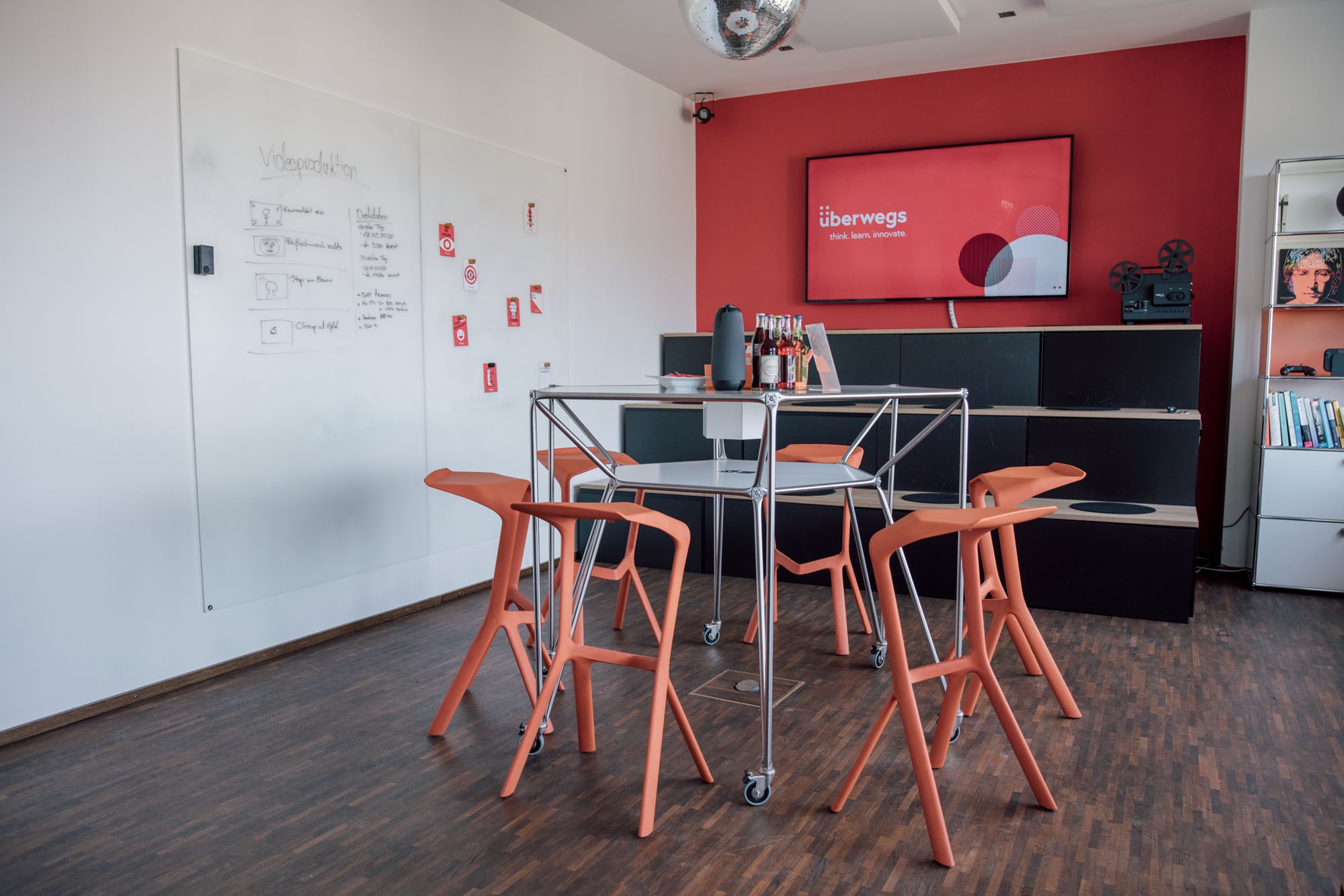 Ein moderner Besprechungsraum mit einem runden Tisch und orangefarbenen Stühlen. An der Wand steht ein Whiteboard mit Notizen zur Videoproduktion. Ein Bildschirm zeigt das Logo "überwegs" und den Slogan "think. learn. innovate