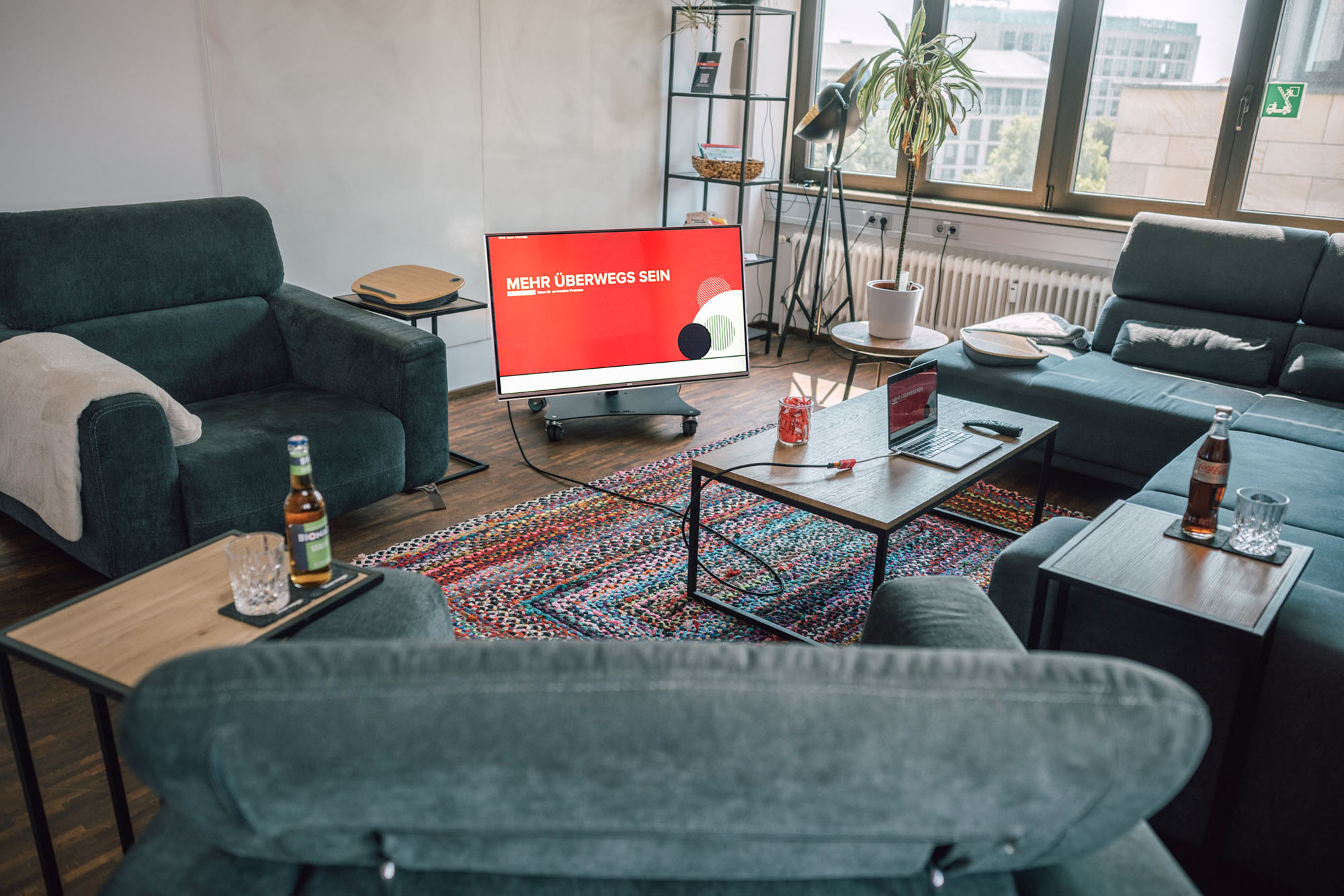 Ein gemütlicher Wohnraum mit zwei grünen Sofas, einem Fernseher, der "MEHR ÜBERWEGS SEIN" anzeigt, und einem Couchtisch mit Getränken